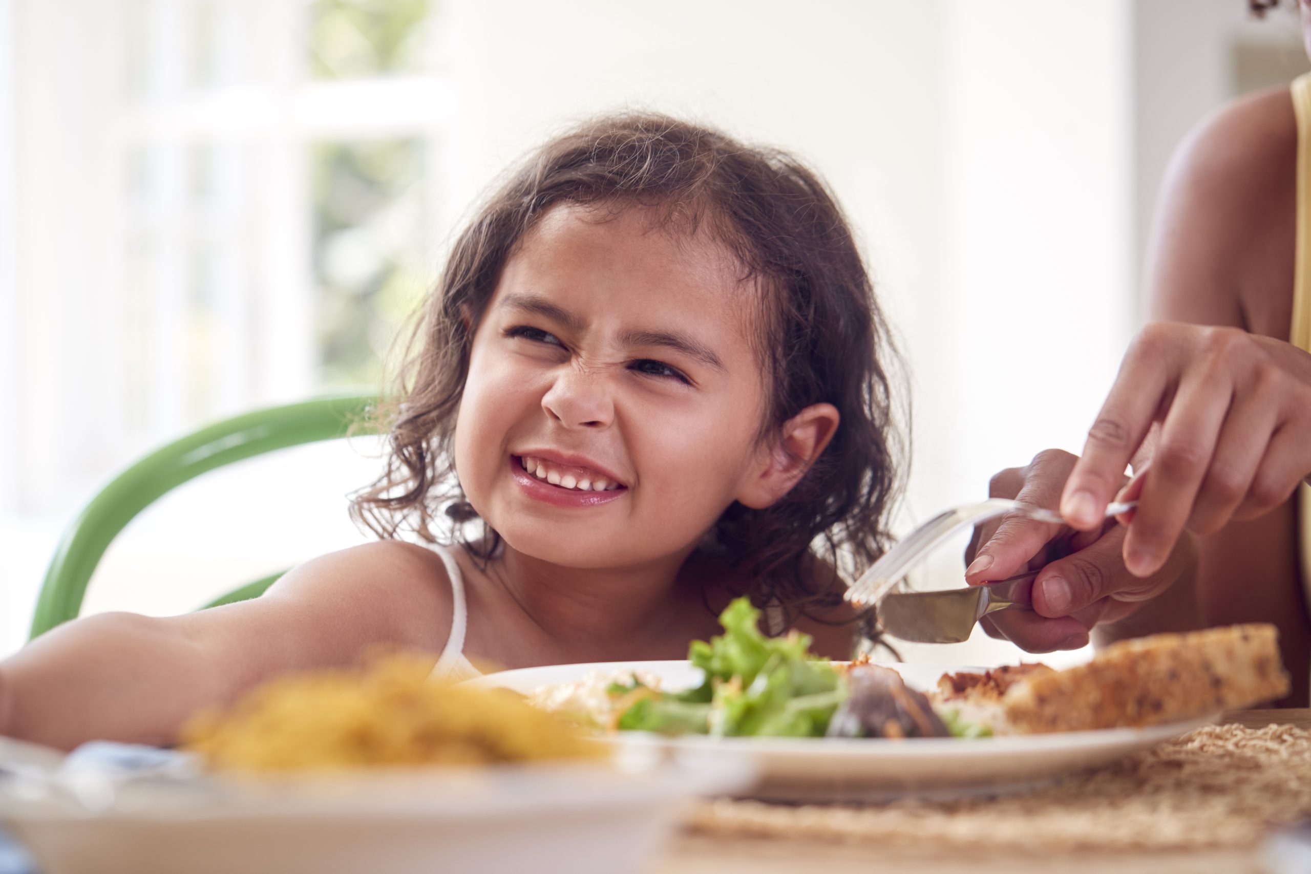 Mother Helping Daughter To Cut Food Sitting Around Table At Home Eating Family Meal Together
